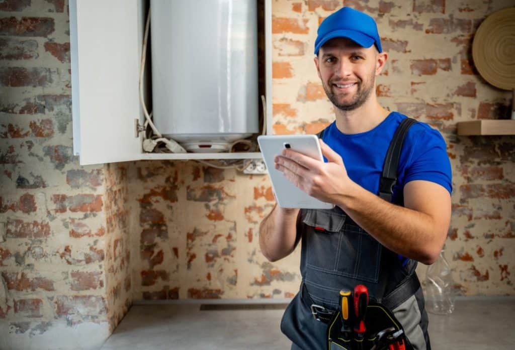 A plumber wearing work overalls and a blue cap holds a tablet beside an open water heater in a brick-walled utility room.