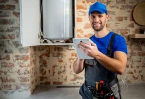 A plumber wearing work overalls and a blue cap holds a tablet beside an open water heater in a brick-walled utility room.