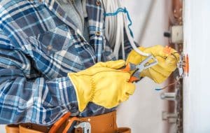 An electrician wearing protective gloves uses wire strippers to repair electrical wiring inside a wall-mounted electrical box.